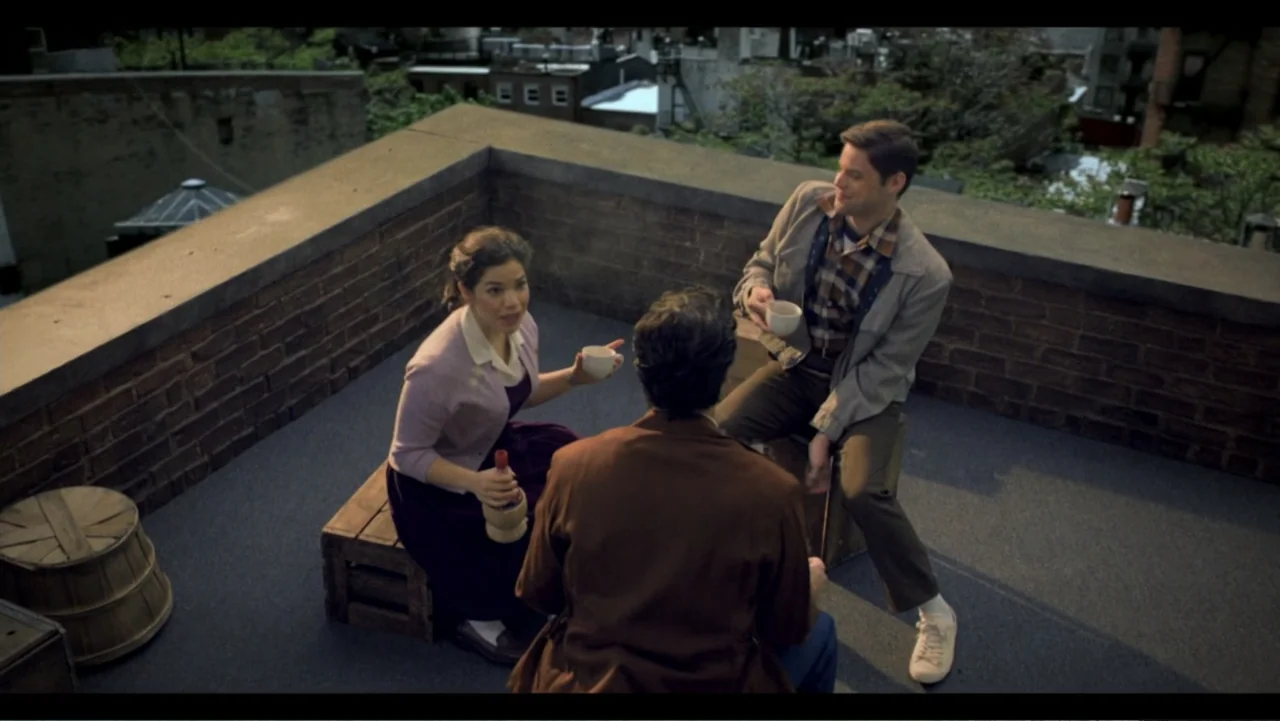 Six by Sondheim (2013) movie still: wide — A brick-walled rooftop terrace overlooks a dense city neighborhood at dusk. Three people sit on wooden…
