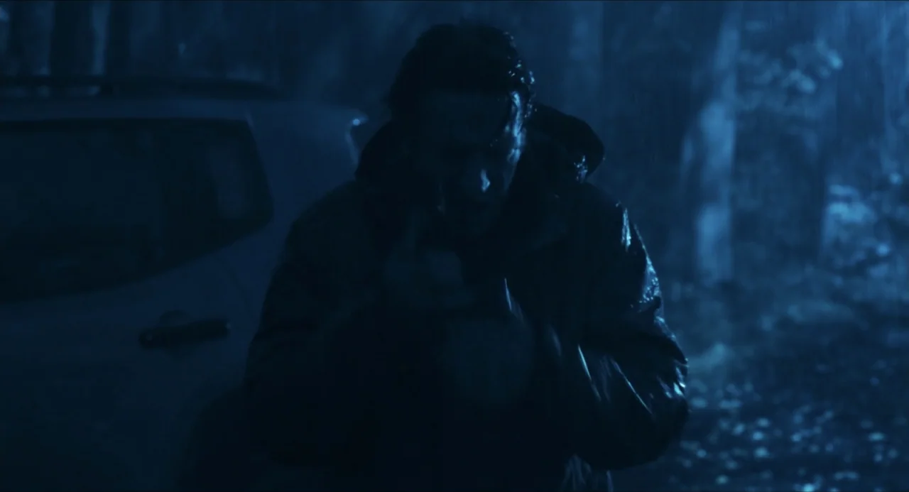 Echoes Of A Crime (2025) movie still: medium — A rain-slicked outdoor environment at night features a dark vehicle parked nearby. A man in a hood…