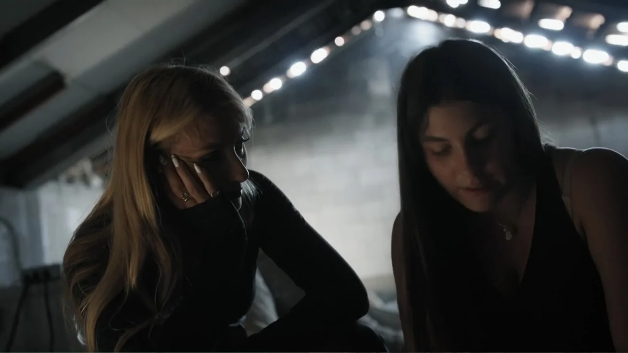 The Children of October 7 (2025) movie still: medium — A dimly lit attic space features low ceilings and a string of hanging lights. Two young women…
