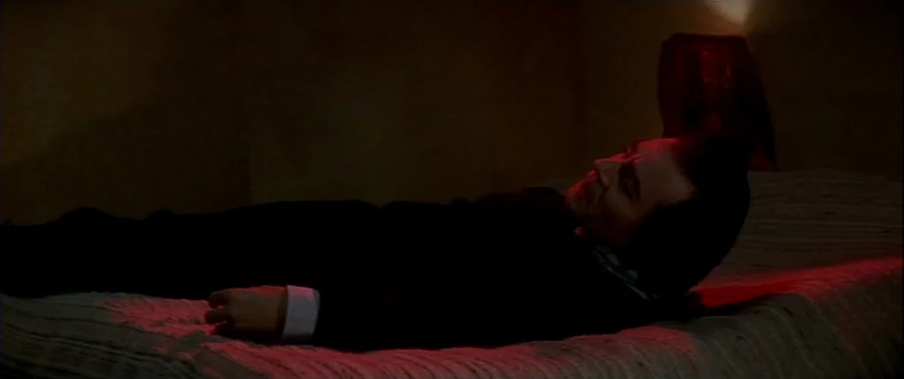 Love Unto Death (1984) movie still: medium — A dark, dimly lit room with minimal furniture and deep red illumination. A man in a dark suit lies flat…
