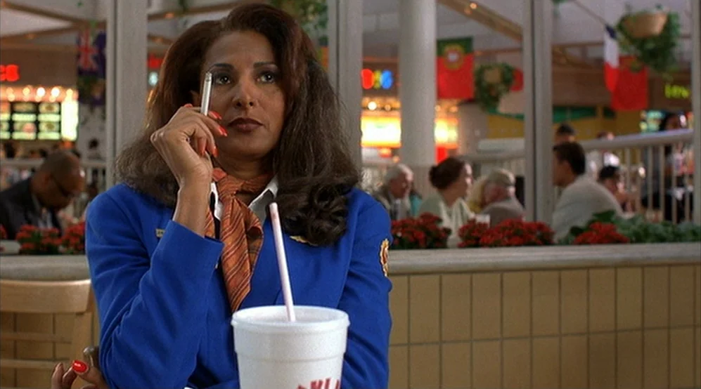 Jackie Brown movie still: medium — A food court seating area features beige tiled partitions and hanging international flags above. An airline…