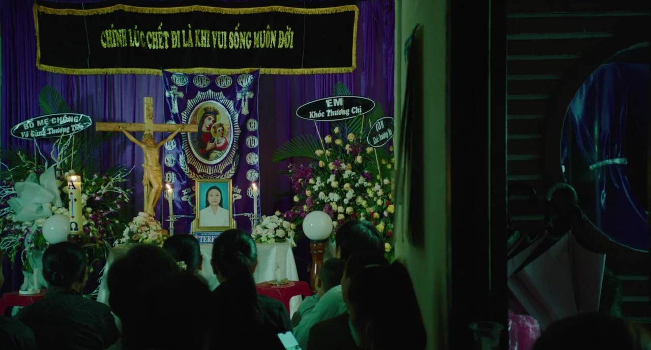 Inside The Yellow Cocoon Shell (2023) movie still: medium — A dimly lit room holds a Catholic funeral altar decorated with purple fabric and…