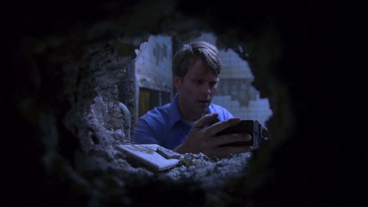 Saw movie still: medium — A man peers through a jagged, irregular hole broken into a tiled interior wall. He holds a small, rectangular black object…