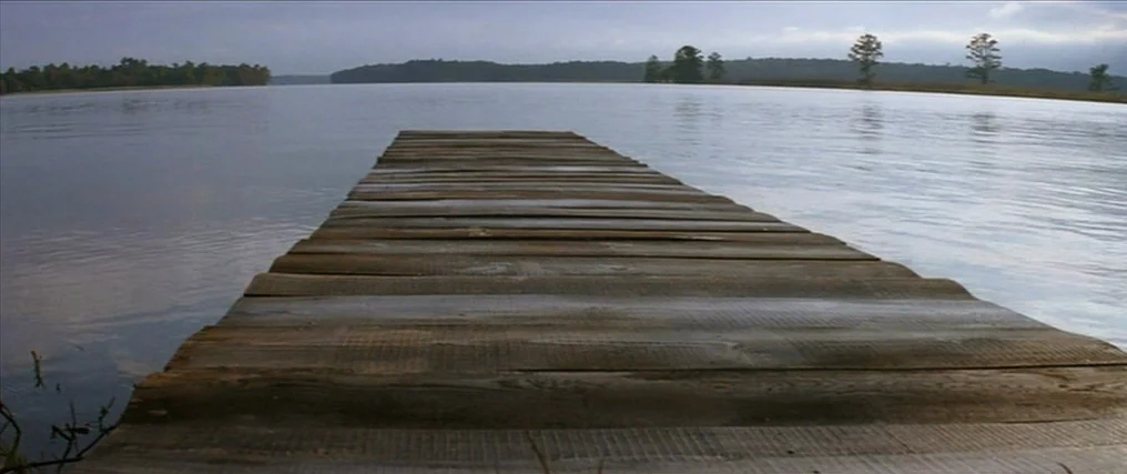 The New World (2005) movie still: establishing — A wooden pier stretches out into the center of a wide, calm lake. The horizon shows a tree line…