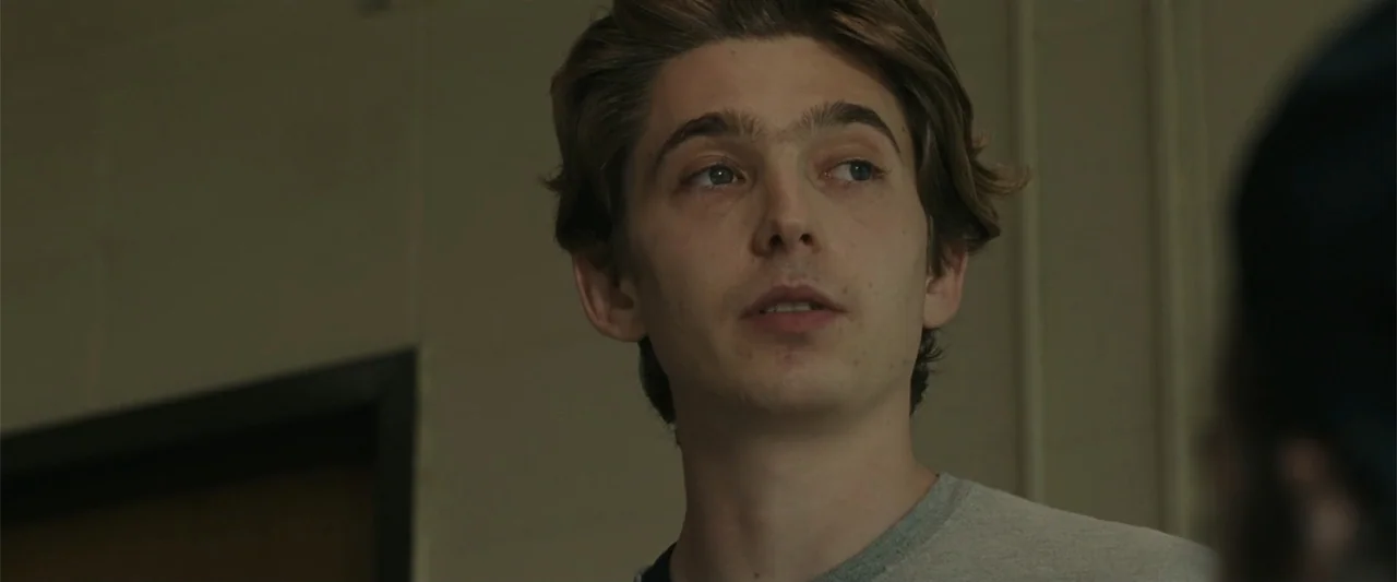 Chemical Hearts (2020) movie still: closeup — A neutrally colored interior space with light-colored walls and a dark door frame. A young man with…