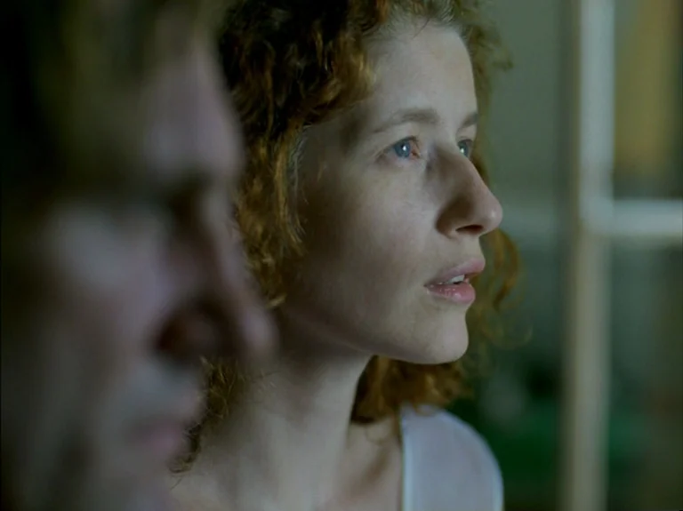 Helas Pour Moi (1993) movie still: closeup — A dimly lit indoor space with muted tones and blurred background elements. A woman with curly red hair…