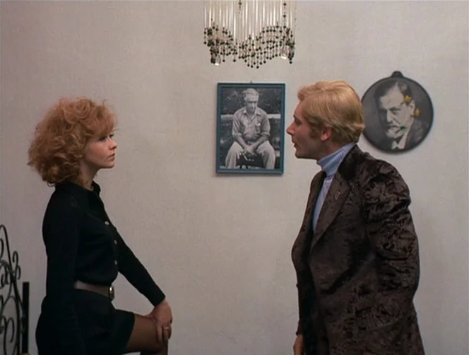 WR: Mysteries of the Organism (1971) movie still: medium — A white-walled room features a crystal chandelier and framed portraits on the wall. A man…