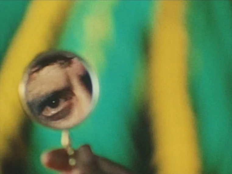 Salome (1972) movie still: closeup — A close-up view shows a hand holding a magnifying glass against a backdrop. The lens displays a reflection of a…