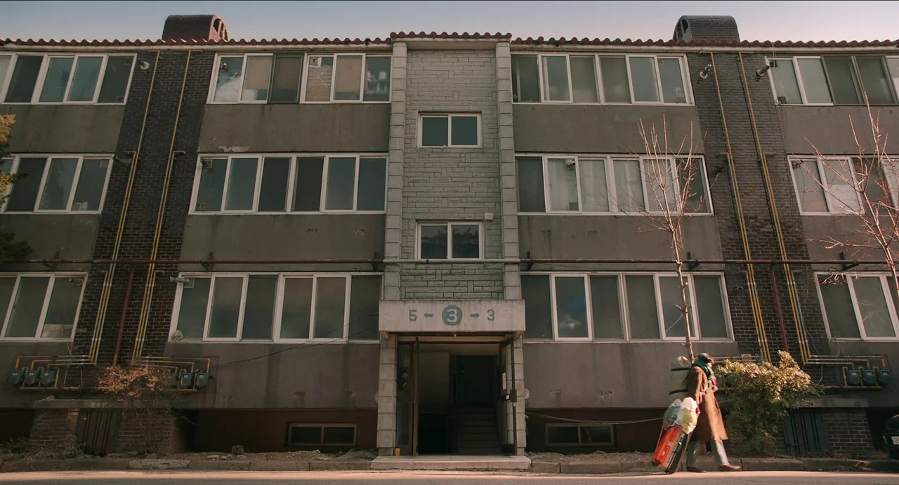 Microhabitat (2017) movie still: wide — A utilitarian four-story apartment complex features grey concrete walls and numerous white-framed windows. A…
