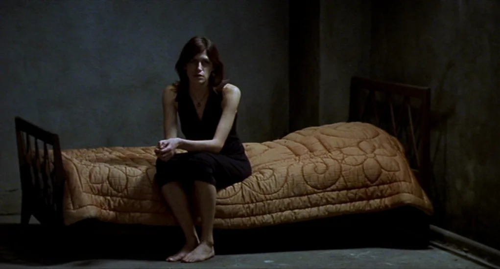 Tiresia (2003) movie still: medium — A dimly lit room with plain gray walls contains a single bed. A woman in a black dress sits on the bed with…