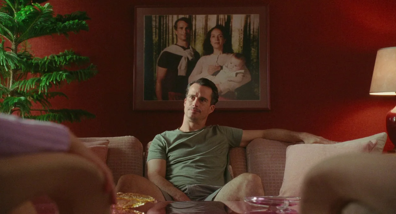 Babysitter (2022) movie still: medium — A man sits on a sofa against a deep red wall with a framed family portrait. He gazes off-camera while…