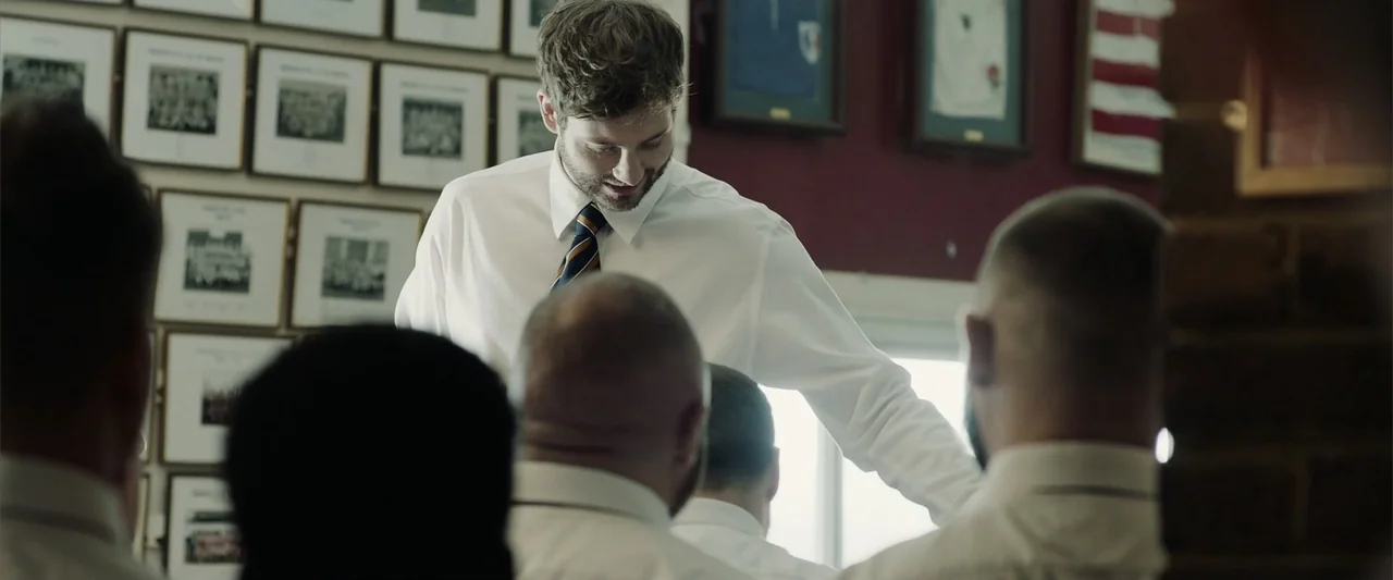 The Thorn On His Side (2014) movie still: medium — A dimly lit club room features walls covered in framed team photographs. A man in a white dress…