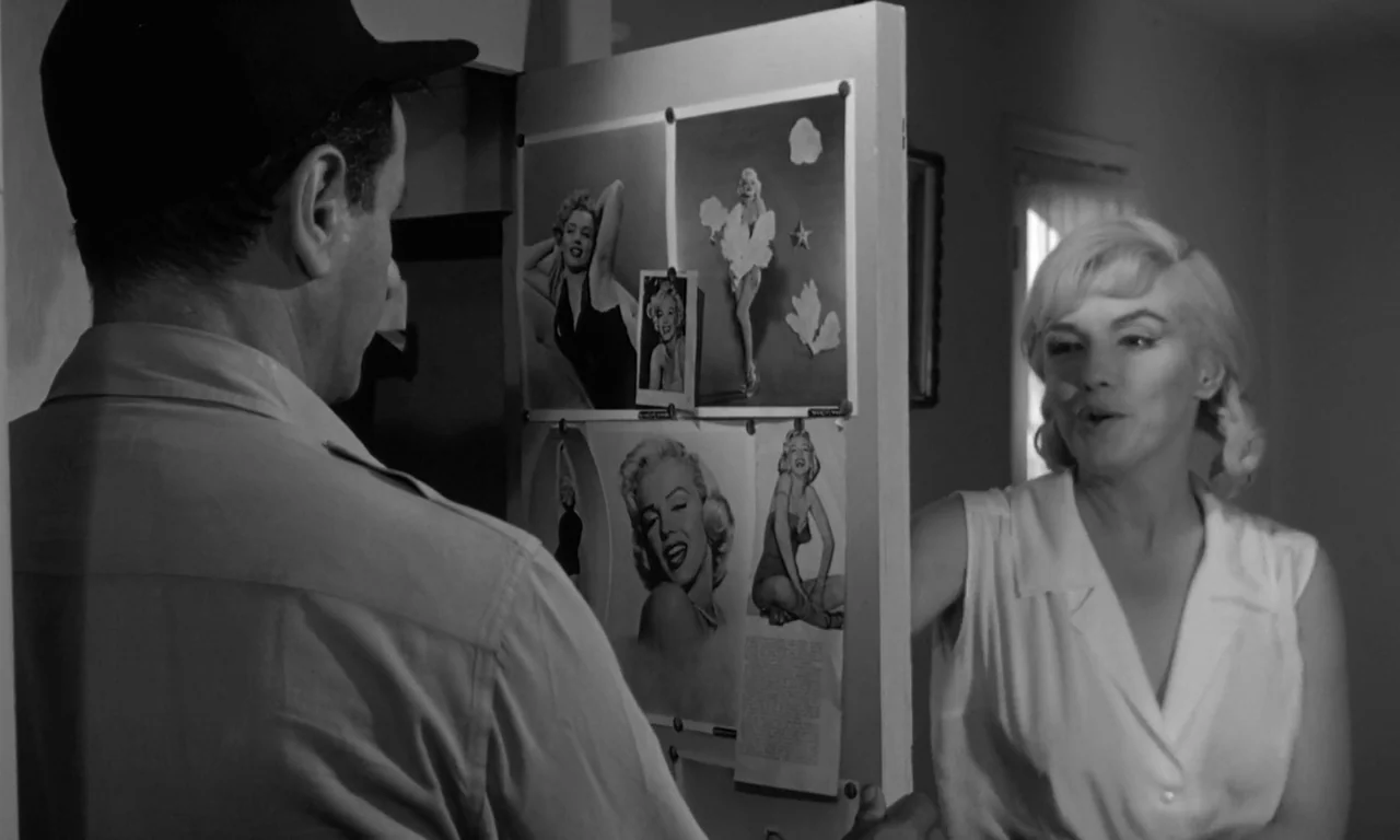 The Misfits (1961) movie still: medium — An interior space features a white display board covered with multiple photographs of a woman. A man and a…