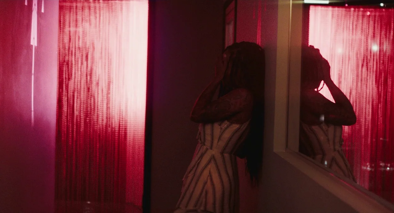Test Pattern (2019) movie still: medium — A dimly lit narrow corridor features a shimmering red beaded partition against the wall. A woman stands…