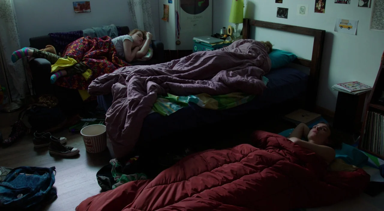 Laggies (2014) movie still: wide — A cluttered room contains three people sleeping on a couch, a bed, and the floor. The space is filled with…