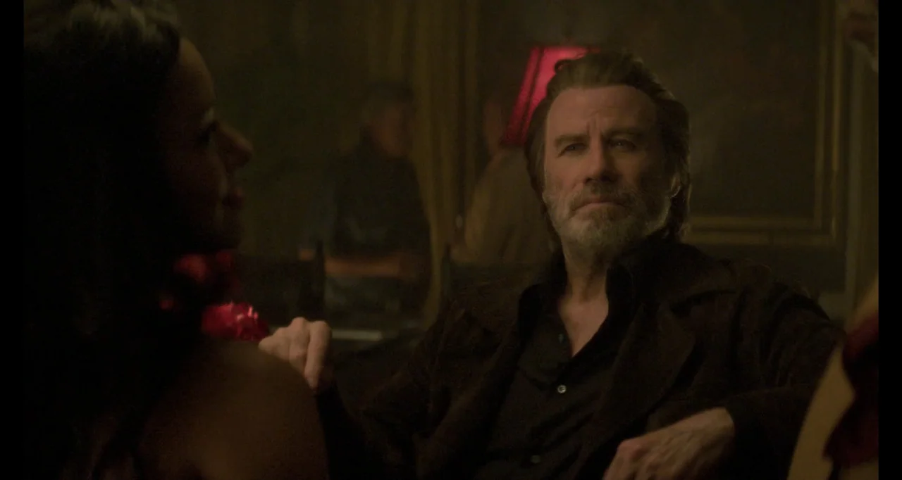 The Poison Rose (2019) movie still: medium — A dimly lit lounge space features dark wall panels and a red-shaded lamp. A middle-aged man sits in the…