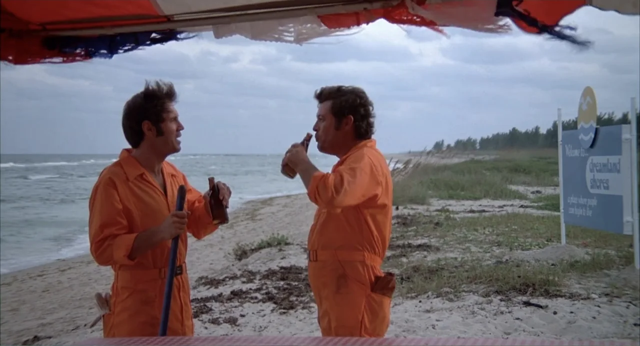 Empire of the Ants (1977) movie still: medium — A sandy beach features a weathered orange canopy and a blue advertisement sign. Two men in bright…