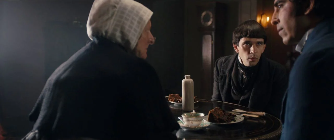 The Personal History of David Copperfield (2019) movie still: medium — Three individuals sit at a circular dark wood table within a dimly lit room.…