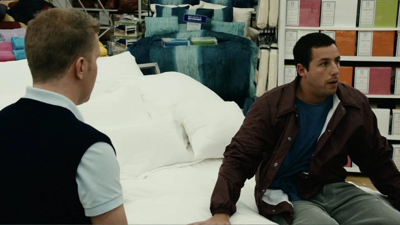 Click (2006) movie still: medium — A retail bedding section displays white pillows and blue bedding in a store aisle. Two men sit on a mattress…