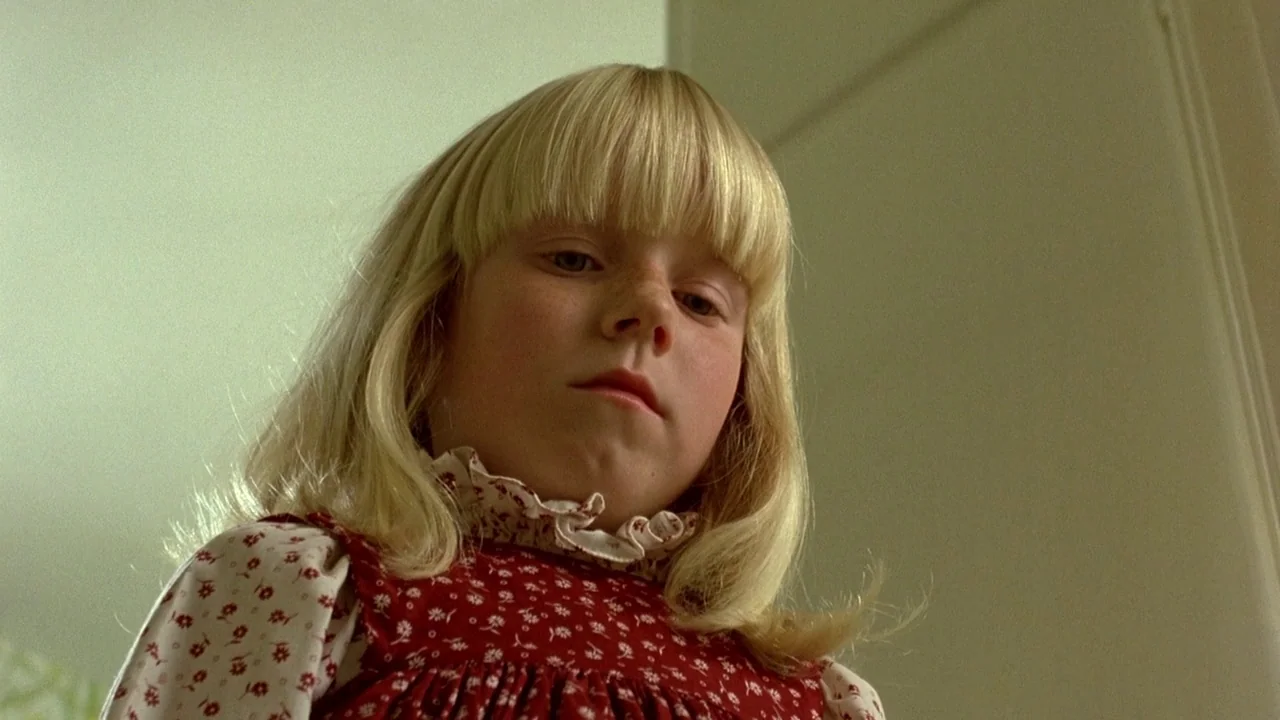 The Brood (1979) movie still: closeup — A plain interior room with light-colored walls serves as the backdrop. A young girl with blonde hair and a…