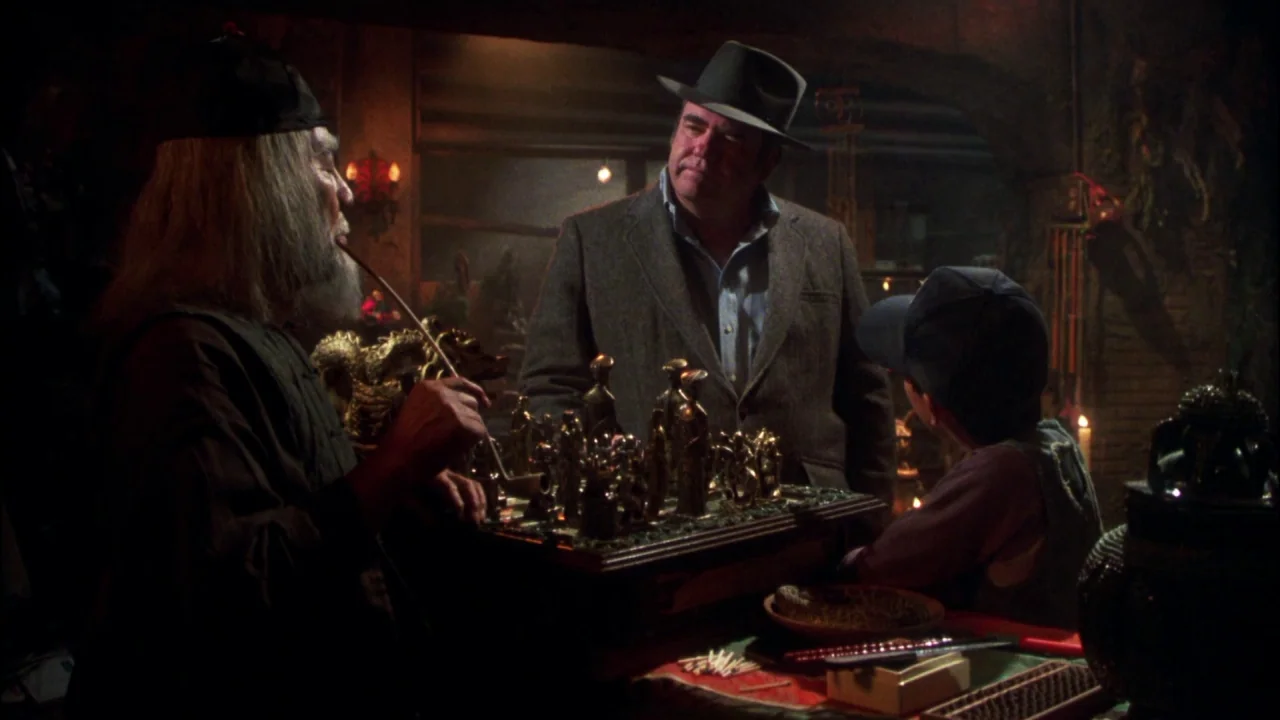 Gremlins (1984) movie still: medium — A dimly lit antique shop contains shelves of cluttered artifacts and a central game board. An elderly man with…