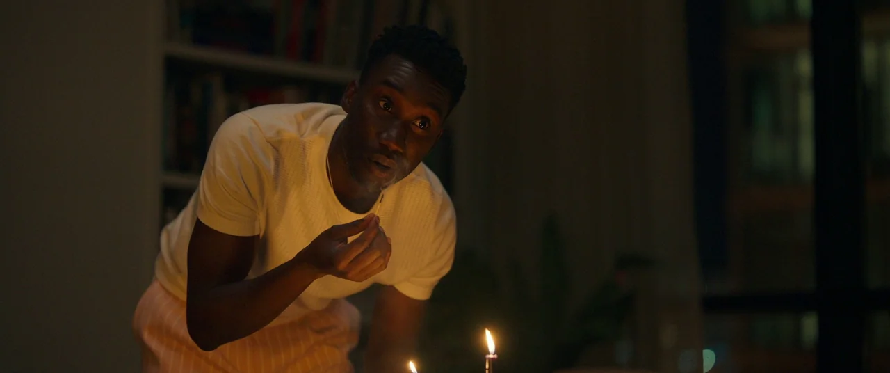 Candyman (2021) movie still: medium — A dark apartment interior at night with blurred bookshelves visible in the background. A man leans over a…