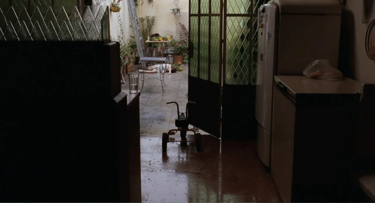 La Ciénaga (2001) movie still: wide — A tiled courtyard patio is visible through an open glass-paned door from a kitchen. A small child lies face…