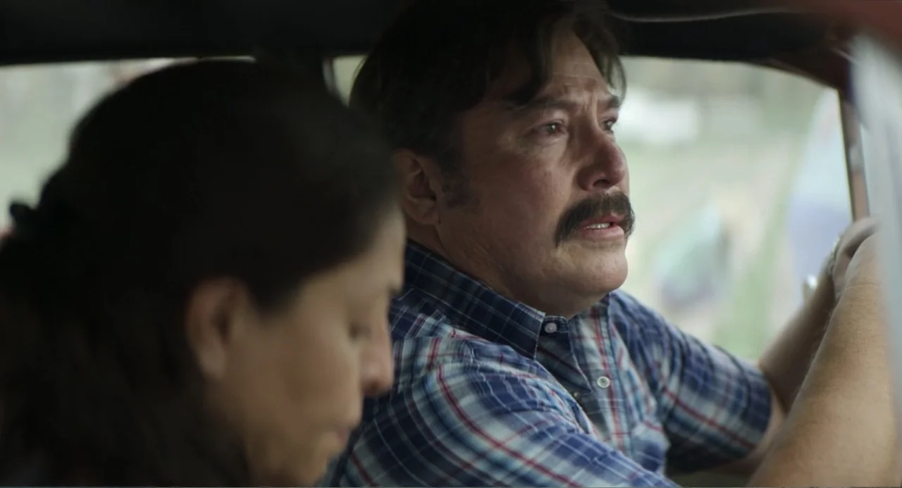 A Million Miles Away (2023) movie still: medium — A car interior during the day with two people seated in the front. A man driving the car expresses…