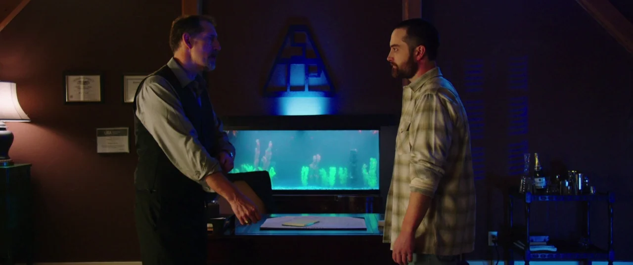 The Mind’s Eye (2015) movie still: medium — A dimly lit room with a desk, an aquarium, and wall art in the background. Two men stand on opposite…