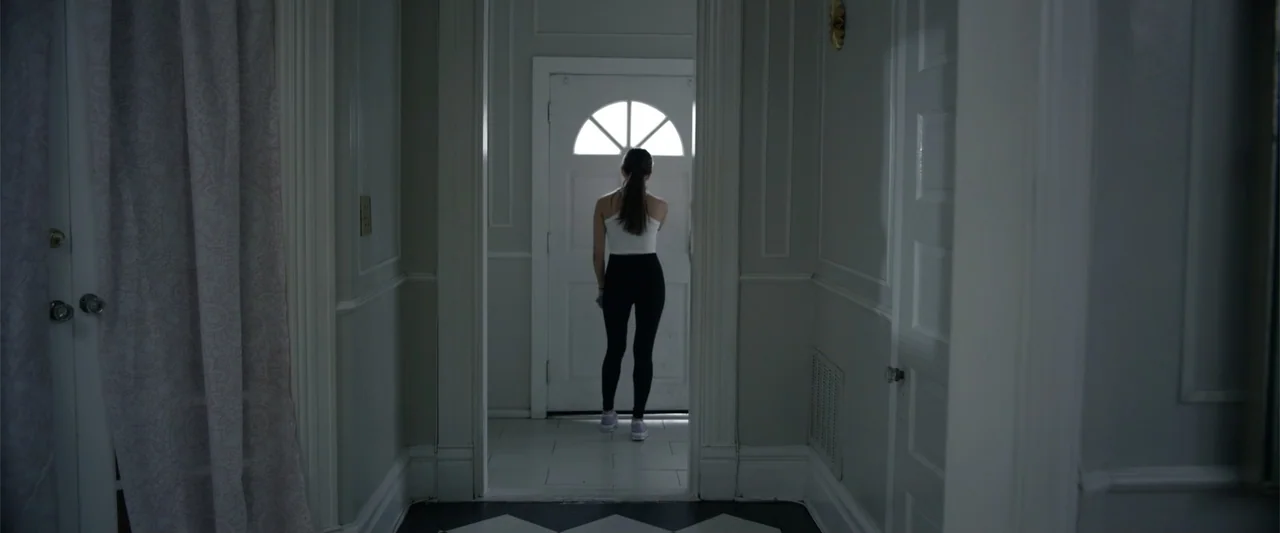 Init!ation (2021) movie still: fullbody — A residential hallway with white walls and a black and white tiled floor. A young woman in athletic wear…
