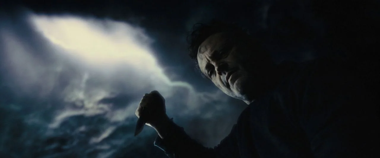 Freaky (2020) movie still: closeup — A dark rocky cavern interior with a bright light source illuminating one side. A man holds a knife up while…
