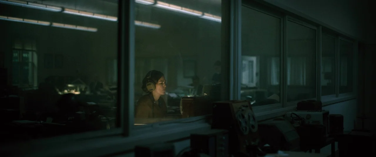 Boston Strangler (2023) movie still: medium — A dimly lit technical room houses a woman seated behind a large glass partition. Equipment including…