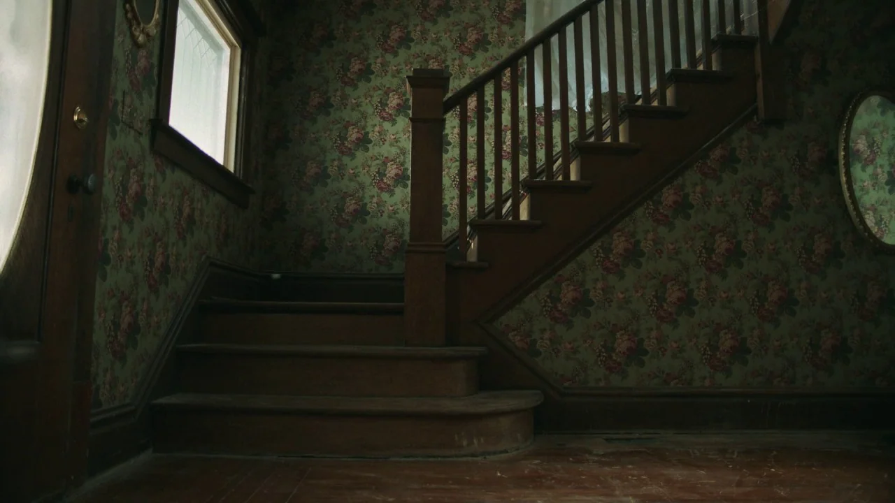 Girl on the Third Floor (2019) movie still: wide — A residential entry with a wooden staircase and walls covered in dark floral wallpaper. The scene…