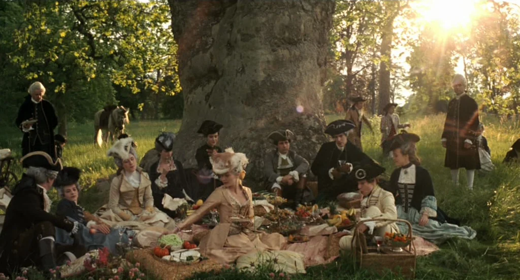 Marie Antoinette (2006) movie still: wide — A group of people in 18th-century attire sit on blankets picnicking under a giant tree. Lush green grass…