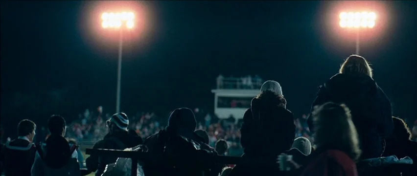 Snow Angels (2007) movie still: wide — A nighttime view from behind spectators sitting in the bleachers of an outdoor sports stadium. Two large,…