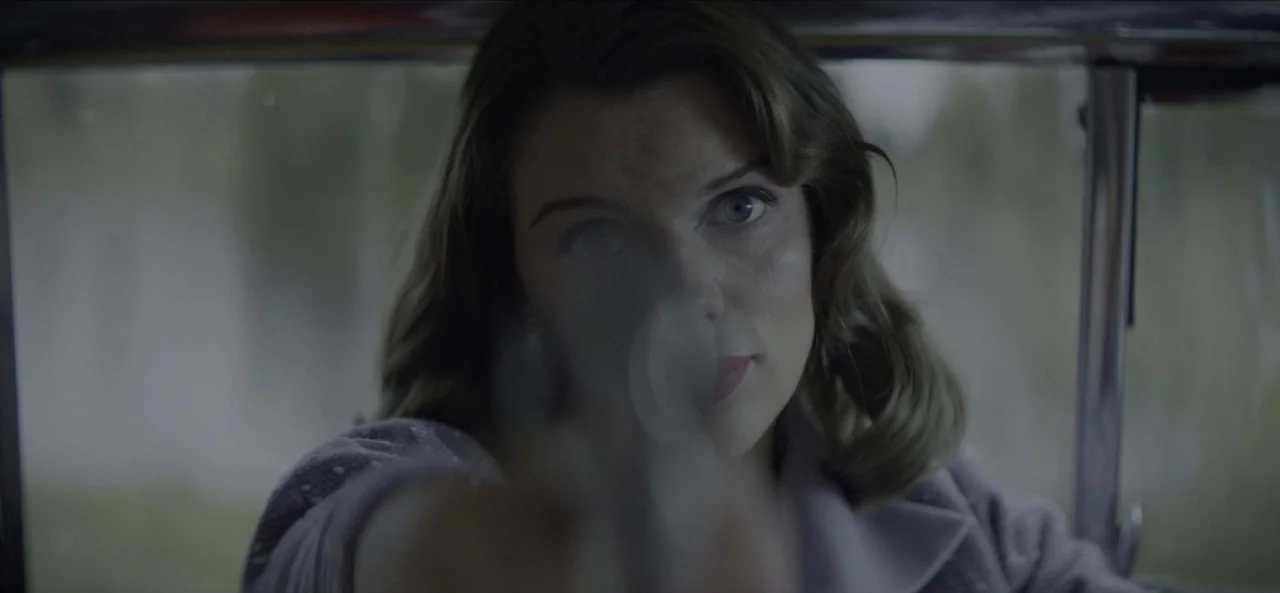No Sudden Move (2021) movie still: closeup — A woman sits inside a car looking through the windshield with a focused gaze. She holds a gun aimed…