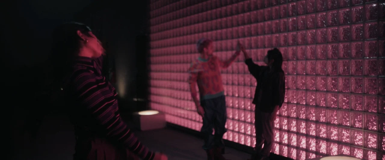 Sign Here (2017) movie still: medium — A dimly lit room features a large, backlit pink glass block wall. Two individuals stand before the wall,…