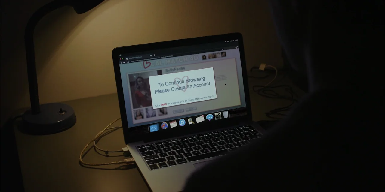 Lobos (2019) movie still: medium — A dark, dimly lit indoor space featuring a desk with a computer and lamp. A person viewed from behind observes a…