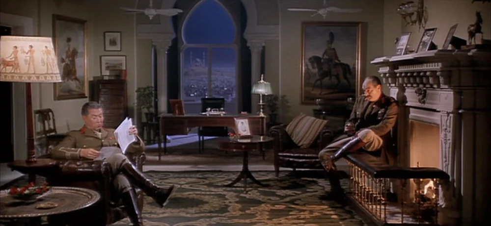 Lawrence Of Arabia movie still: wide — A formal study features ornate rugs, leather armchairs, and a large fireplace with glowing flames. Through a…