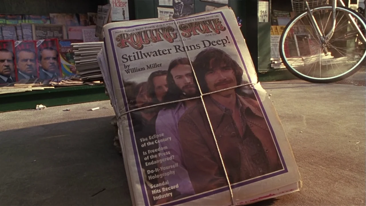 Almost Famous (2000) movie still: closeup — A stack of tied Rolling Stone magazines sits on a concrete sidewalk near a newsstand. The cover features…