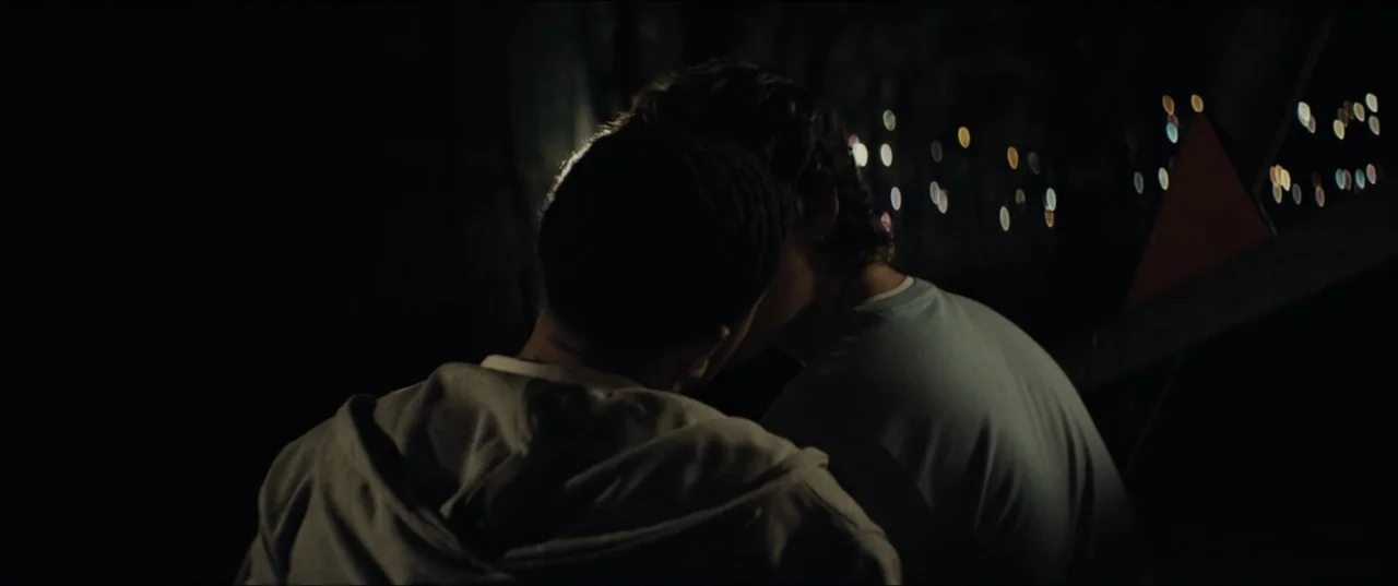 Bonus Track (2024) movie still: medium — A dark outdoor setting at night features blurred bokeh lights in the distance. Two figures lean toward each…