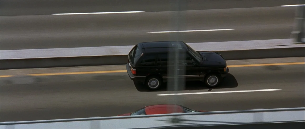 Igby Goes Down (2002) movie still: wide — An asphalt multi-lane highway stretches across the frame during bright daylight conditions. A dark SUV…