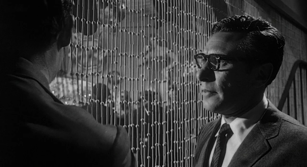 Touch of Evil (1958) movie still: medium — A dimly lit room is divided by a hanging string curtain with round beads. A man in a suit wearing glasses…