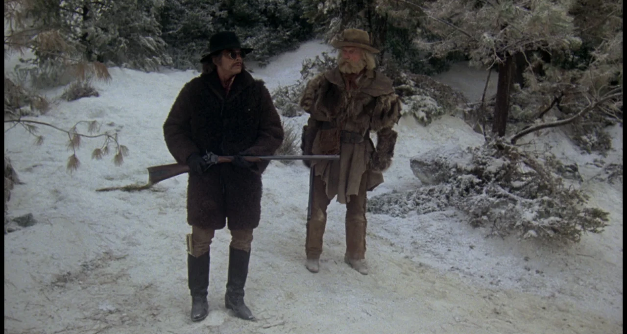 The White Buffalo (1977) movie still: fullbody — A snow-covered clearing in a forest with tall pine trees in the background. Two men dressed in…