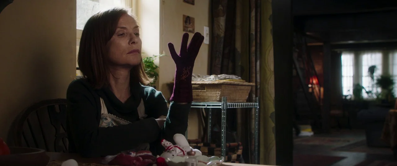 Greta movie still: medium — A cluttered room features a wooden table and a metallic wire shelving unit nearby. An older woman sits at the table,…