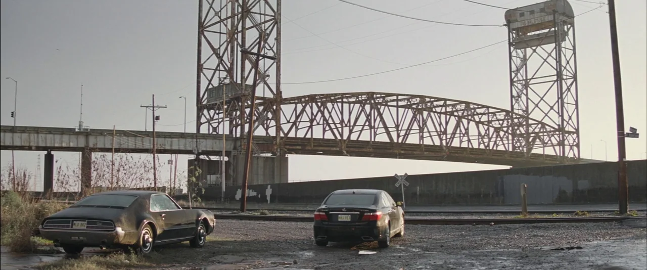 Killing Them Softly (2012) movie still: wide — A gravel lot sits beneath a massive, rusting metal lift bridge during daytime. Two black automobiles…