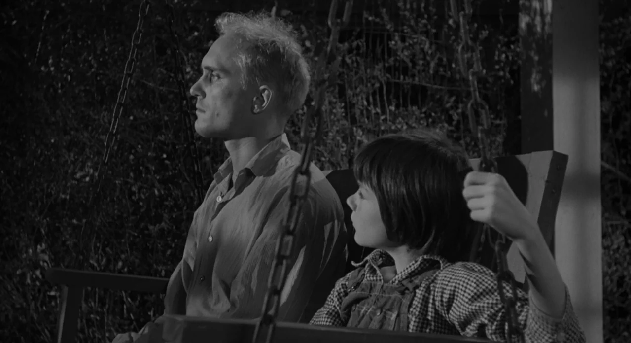 To Kill A Mockingbird (1962) movie still: medium — A man and a boy sit together on a porch swing at night. Dark, dense foliage serves as the…