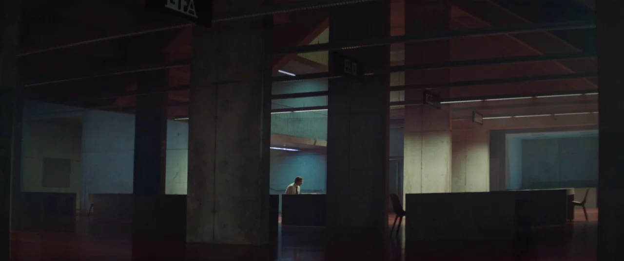 The Platform (2019) movie still: wide — A dark, sprawling modern interior with exposed concrete pillars and minimalist workstation dividers. A lone…