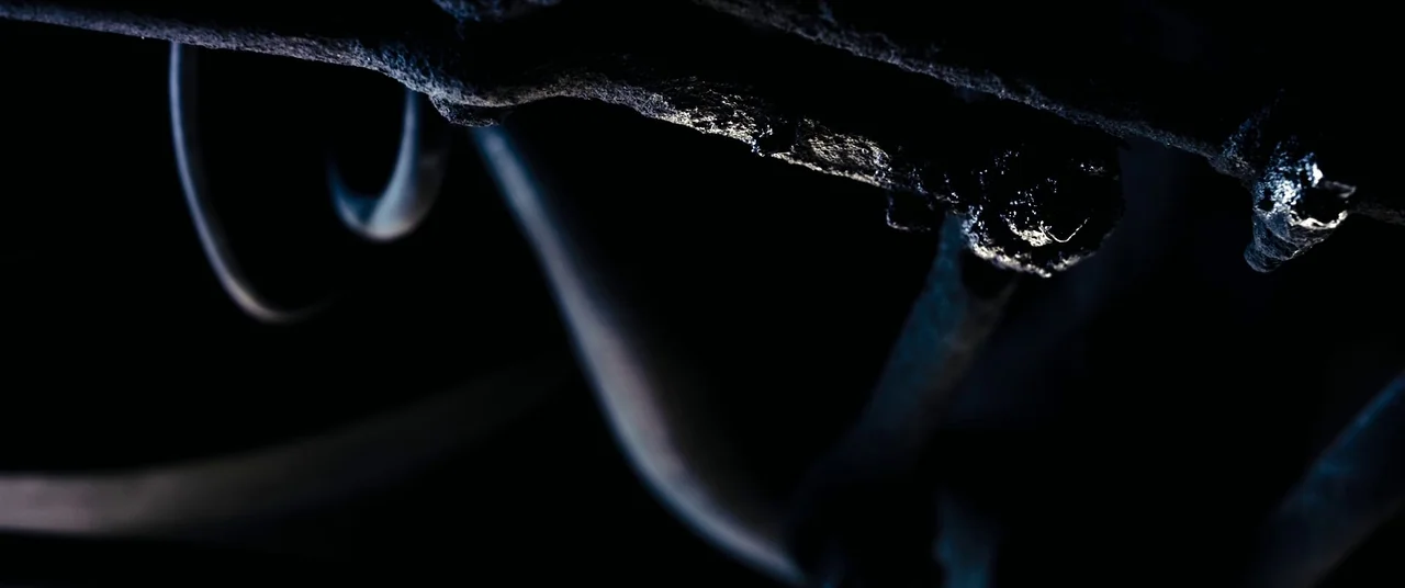 Titane (2021) movie still: closeup — Close-up detail of rusted, metallic mechanical parts viewed from beneath an automobile. Dark surfaces catch a…