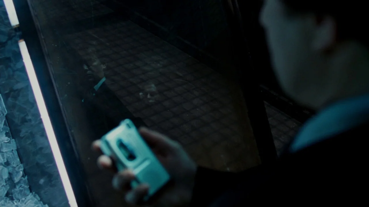 Saw V (2008) movie still: closeup — A dark, modern space features a reflective glass panel lit by long, vertical lights. A man stands out of focus…